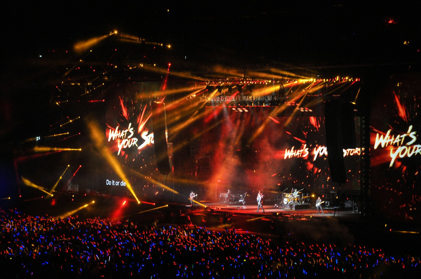 Una band si esibisce sul palco davanti a una folla che sventola bastoncini luminosi.
